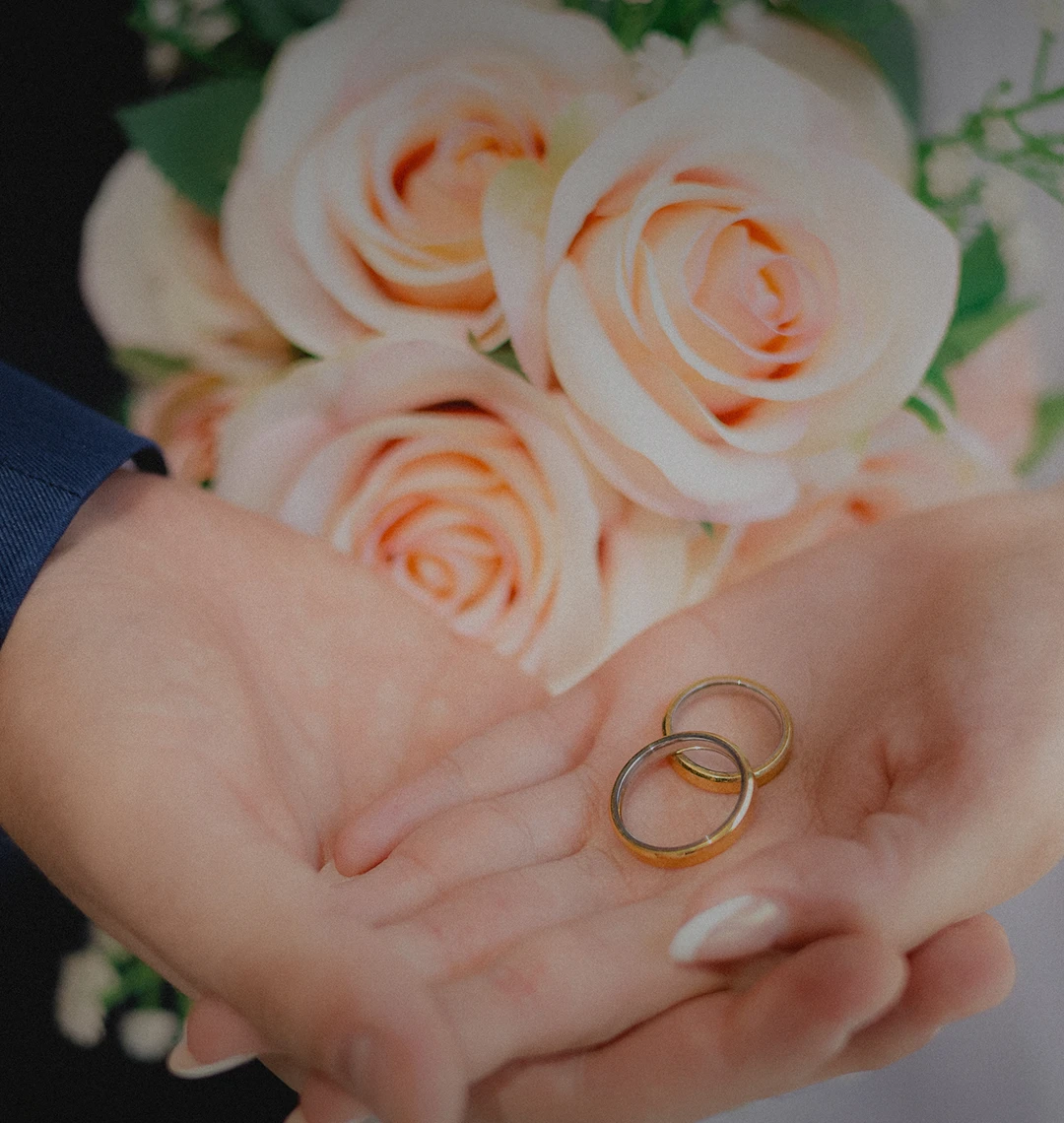 Alianças nas mãos do casal, em frente a um buquê de rosas claras